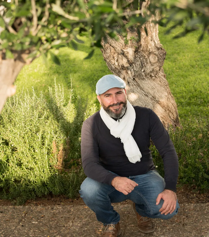 Antoine Hadjialexandrou, garden designer in Paphos, crouching among rosemary beneath an old olive tree