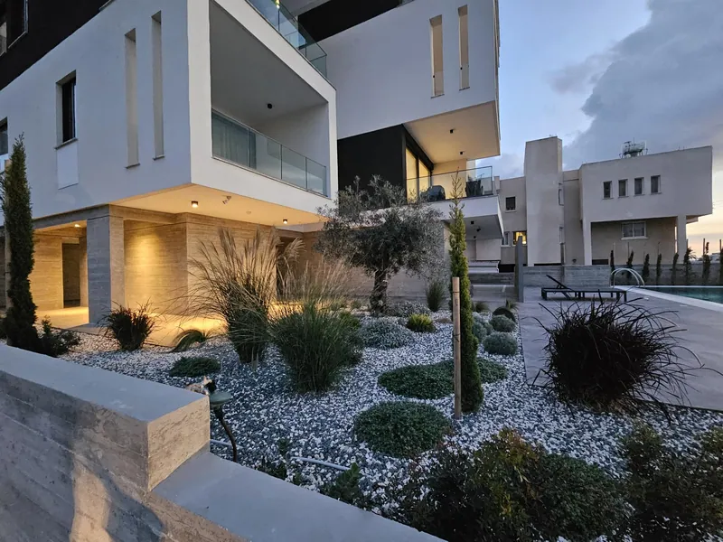 Grasses, shrubs and a young cypress in white gravel beside the building, lit at dusk