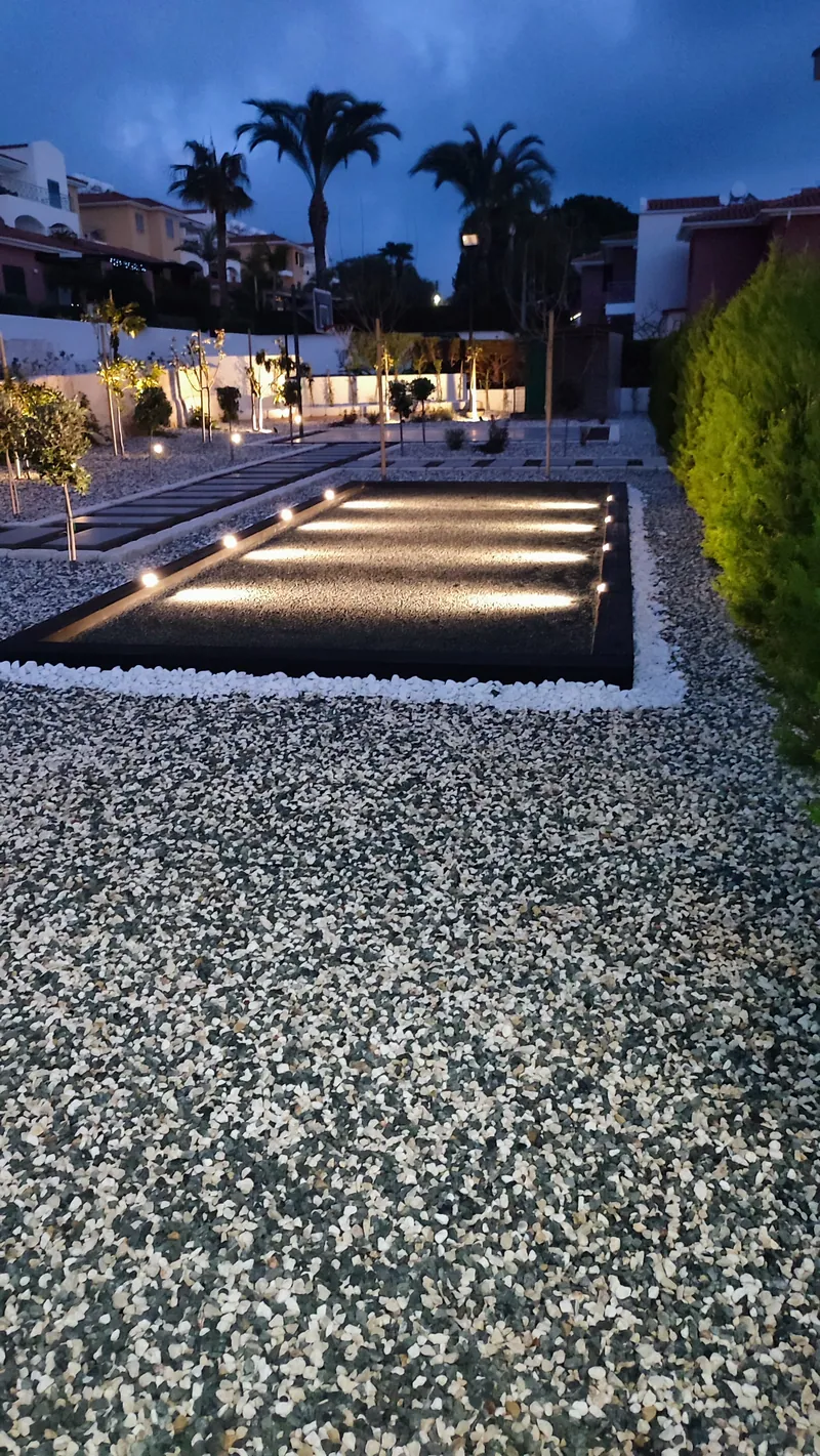 A lit reflecting pool at dusk in a garden of speckled gravel, palm trees beyond