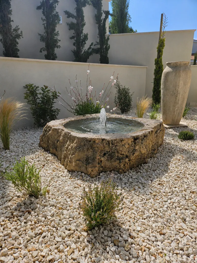A bubbling stone basin in white gravel beside a tall clay jar, with young grasses and shrubs